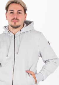 Lichtgrijze zip-up hoodie met een trekkoordkap, voorzakken en geribbelde manchetten. Beschikt over een klein logo op de linkermouw.