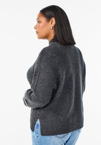 Grijze gebreide trui met een hoge hals, verlaagde schouders, zijsplitten en ribgebreide manchetten, gecombineerd met lichtblauwe jeans.