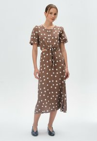 Braun gepunktetes Kleid mit gebundenem Bund, kurzen Puffärmeln und Midi-Länge. Der Stoff wirkt glatt und leicht; kombiniert mit navyfarbenen Schuhen.