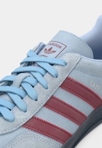 Ανοιχτό μπλε αθλητικό παπούτσι Adidas Gazelle με κόκκινες ρίγες και κορδόνια σε ανοιχτό μπλε χρώμα, δείχνοντας λεπτομερή ραφή και λογότυπο στη γλώσσα και στο πλάι.
