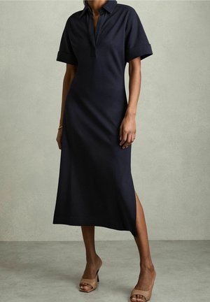 Frau trägt ein marineblaues Midikleid im Polostil mit kurzen Ärmeln und einem Seitenschlitz, kombiniert mit beigen, offenen Sandalen mit hohem Absatz.