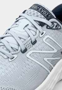 Light gray athletic shoe with a textured knit upper, white synthetic accents, and flat laces. Features a padded collar and logo on the tongue.