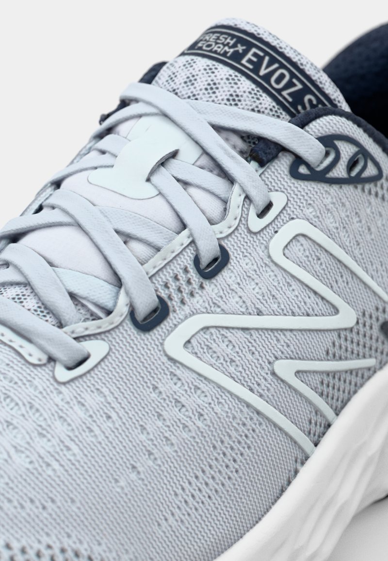Zapatilla deportiva de color gris claro con parte superior de punto texturizado, acentos sintéticos en blanco y cordones planos. Presenta un collar acolchado y un logotipo en la lengüeta.