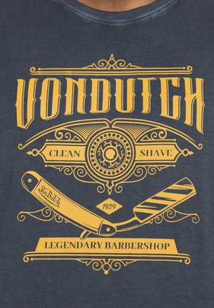 Camiseta de color navy con un gráfico amarillo que incluye una navaja recta, espirales decorativas y el texto: "VON DUTCH, AFEITADO LIMPIO, BARBERÍA LEGENDARIA, 1929."