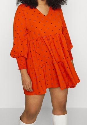 Femme portant une robe ample orange à pois noirs, avec des manches longues bouffantes, et des bottes blanches montant jusqu'aux genoux.