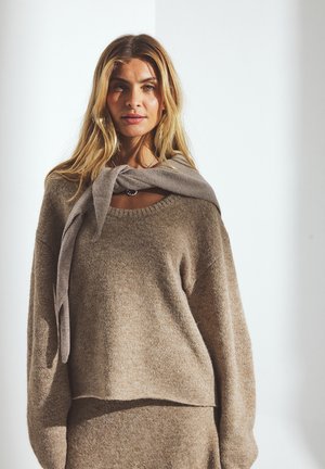 Beige knitted sweater with a rounded neckline and dropped shoulders, paired with a gray scarf tied loosely around the neck. Soft texture.