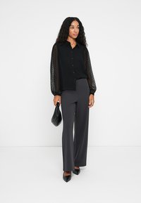 Blouse noire à manches transparentes à pois, avec un boutonnage à l'avant, associée à un pantalon taille haute gris foncé et des escarpins noirs à bout pointu. Portant un sac noir.