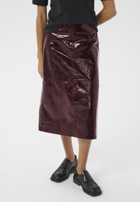 Une personne porte une chemise noire et une jupe midi bordeaux brillante, associée à des chaussures noires épaisses, se tenant devant un fond blanc uni.