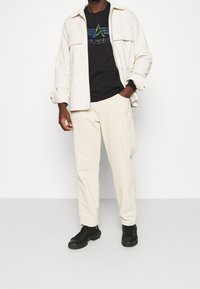 Giacca bianca in corduroy sopra una maglietta nera con logo multicolore, abbinata a pantaloni crema e sneakers nere. Tessuto testurizzato, vestibilità rilassata.