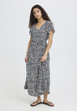 Žena s dugom kovrčavom kosom u crno-bijeloj midi haljini s leopard uzorkom i crnim japankama, stoji na jednobojnoj pozadini.