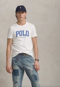 Tricou alb din bumbac cu text albastru "POLO RALPH LAUREN", asortat cu blugi denim uzați cu un design din patchwork și o curea maro.