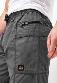 Grå arbejdsshorts med elastisk talje, justerbar snor, sidelomme med lynlås og vævet mærke.