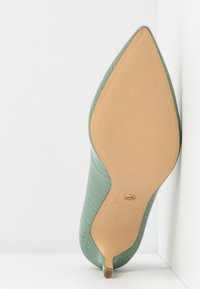 Chaussure à talon haute en cuir synthétique vert effet crocodile avec un bout pointu et une semelle beige, ornée d'un petit accent doré. Taille indiquée : 37.