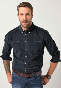 Camisa azul marino de botones con un sutil patrón de cuadros, hecha de tela texturizada. Mangas largas arremangadas, combinada con un cinturón marrón y un reloj.