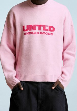 Person trägt einen pinken Pullover mit rotem Schriftzug "UNTLD UNTTLED GOODS" und schwarze Hosen, Hände in den Taschen.
