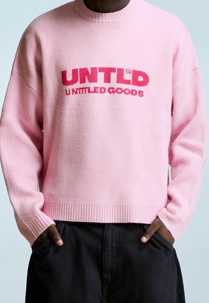 Personne portant un pull rose avec un texte rouge « UNTLD UNTTLED GOODS » et un pantalon noir, mains dans les poches.