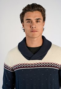 Marineblauwe wollen trui met een crèmekleurig bovenstuk. Heeft een brede sjawlboord, een zigzagpatroon in rood en marineblauw, en kleine witte spikkels door het hele ontwerp.