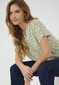 Top verde de manga corta con un patrón abstracto blanco, diseño de escote en V, combinado con jeans de denim azul oscuro. Textura de tela suave.