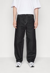 Hombre que lleva puestos unos pantalones vaqueros negros de corte holgado con costuras en contraste blancas y zapatillas deportivas blancas, de pie contra un fondo sencillo.