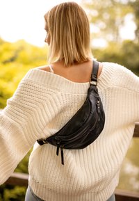 Zwarte leren fanny pack diagonaal gedragen over een witte gebreide trui. Heeft ritsen, een gebogen vorm en een gladde textuur.