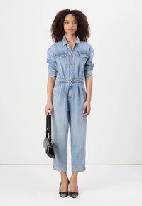 Denim kombinacija z blagim modrim pranjjem, gumbnimi odprtji spredaj, dvema žepoma na prsih, elastičnim pasom in zoženimi nogami. V kombinaciji z črnimi dodatki in petami.