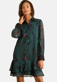 Rochie verde, transparentă, cu un model floral în nuanțe de violet și teal. Dispune de un închidere cu nasturi în față, guler și manșete negre, precum și un tiv cu volane stratificate.