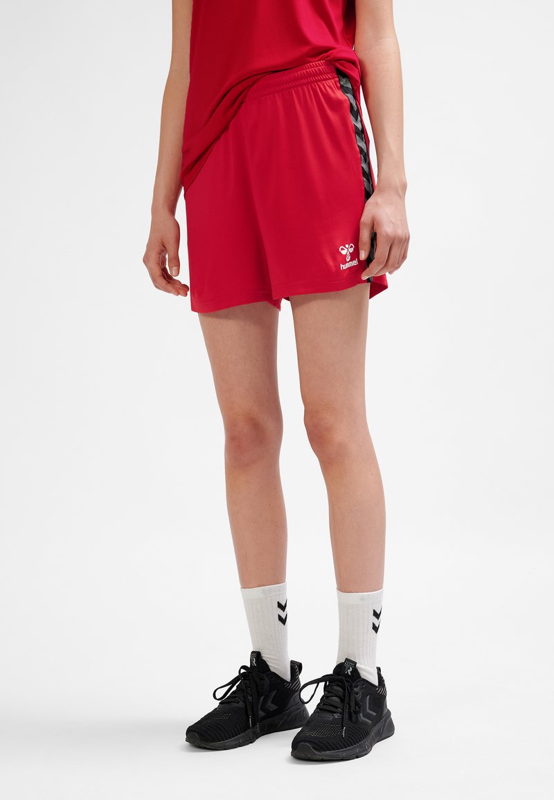 Rode atletische korte broek met zwarte zijstrepen, elastische tailleband en wit logo. Draagt zwarte sneakers en witte sokken met zwarte chevrons.