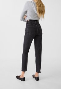 Mujer con cabello largo que lleva jeans negros de tiro alto, sudadera gris de canalé y bailarinas negras, de pie con los brazos cruzados.