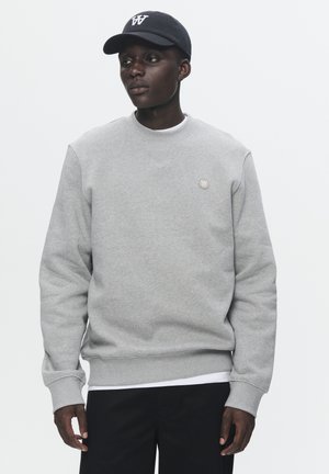 Gray sweatshirt with ribbed cuffs and hem, featuring a small circular logo. Paired with a black cap displaying a white logo. Black pants.