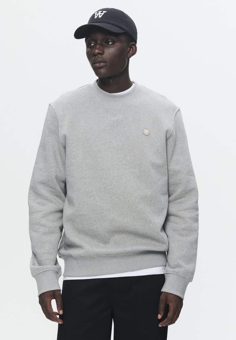Gray sweatshirt with ribbed cuffs and hem, featuring a small circular logo. Paired with a black cap displaying a white logo. Black pants.