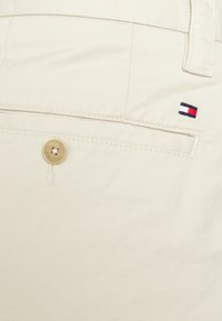 Pantaloni in cotone beige con una tasca posteriore caratterizzata da un piccolo logo ricamato in rosso, bianco e blu, e una chiusura a bottone.