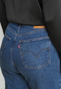 Vaqueros de mezclilla azul con cintura alta, con una etiqueta de cuero en la parte trasera y una etiqueta roja. Diseño estándar de cinco bolsillos con costuras sutiles.