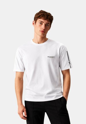 Giovane uomo con capelli ricci indossa una t-shirt bianca Trussardi Swim e pantaloni neri, in piedi con una mano in tasca su uno sfondo semplice.