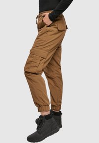 Bruine cargo broek met elastische boorden en zakken aan beide zijden, gedragen met zwarte veterschoenen, wat een casual, functioneel ontwerp benadrukt.