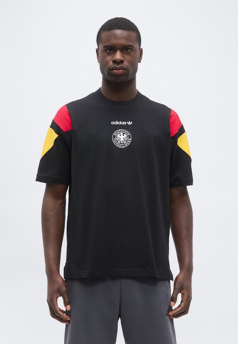 Άντρας με μαύρο t-shirt Adidas με κόκκινα και κίτρινα πάνελ στους ώμους και το έμβλημα της Γερμανικής Ποδοσφαιρικής Ομοσπονδίας, στέκεται μπροστά σε απλό φόντο.
