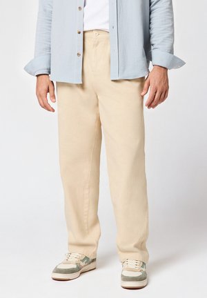 Homme portant un pantalon chino beige, des baskets blanches avec des accents verts, une chemise blanche et une chemise boutonnée bleu clair à rayures avec les manches retroussées.