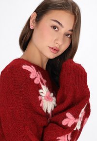 Roter gestrickter Pullover mit floraler Stickerei in Rosa und Weiß, mit lockerer Passform und strukturiertem Stoff. Das Modell trägt blumenförmige Ohrringe.