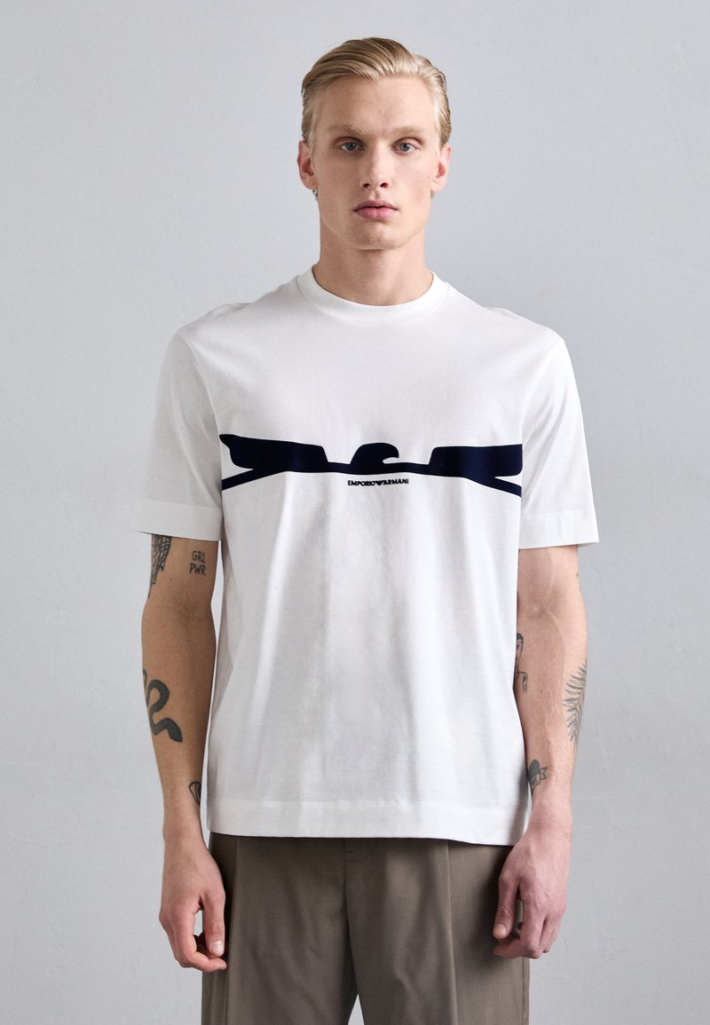 Witte t-shirt van katoen, met een marineblauw grafisch ontwerp en logo op de borst. Korte mouwen en ronde hals.