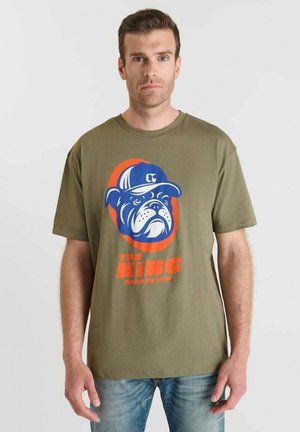 Homme portant un t-shirt vert olive avec un bulldog cartoon bleu et orange portant une casquette, et le texte orange "LE ROI", se tenant contre un fond uni.