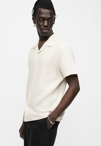 Άνδρας με σκούρο δέρμα και dreadlocks, φοράει κοντομάνικο κρεμ πουκάμισο με κουμπιά και μαύρο παντελόνι, κοιτάζει προς τα πλάγια με το ένα χέρι στην τσέπη.