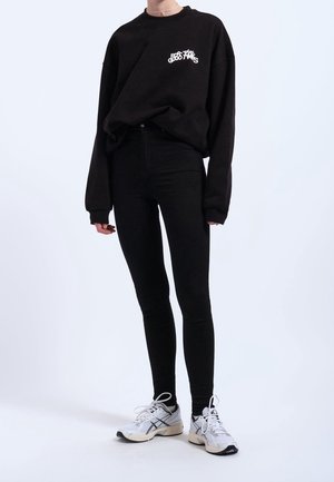 Osoba w czarnej, luźnej bluzie z białym napisem, czarnych dopasowanych spodniach, białych sneakersach, stojąca na jasnym tle.