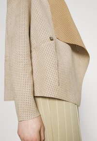 Haut beige perforé à coupe décontractée, avec un accent marron contrastant sur l'épaule et un détail minimaliste en métal argenté.