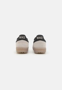 Ténis Nike com esquema de cores em preto e bege, material em camurça, forma arredondada e sola de borracha. Logótipo "NIKE" exibido em destaque no calcanhar.