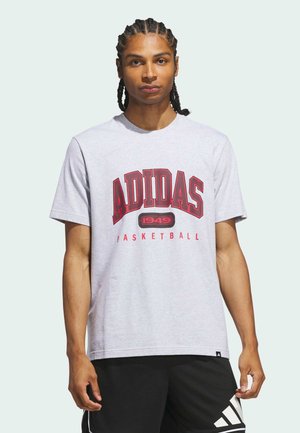Młody mężczyzna z zaplecionymi włosami, ubrany w jasnoszary koszulkę do koszykówki Adidas z czerwonymi napisami i czarne spodenki Adidas.
