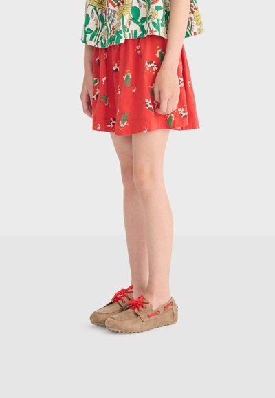 Enfant portant une jupe rouge avec un imprimé d'animaux de la ferme et de fermier, des mocassins beiges avec des lacets rouge vif, et un haut à motif floral.