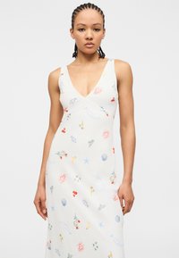 Νεαρή γυναίκα με πλεξούδες φοράει αμάνικο λευκό φόρεμα με βαθύ ντεκολτέ και μικρά πολύχρωμα μοτίβα με θαλασσινό θέμα.