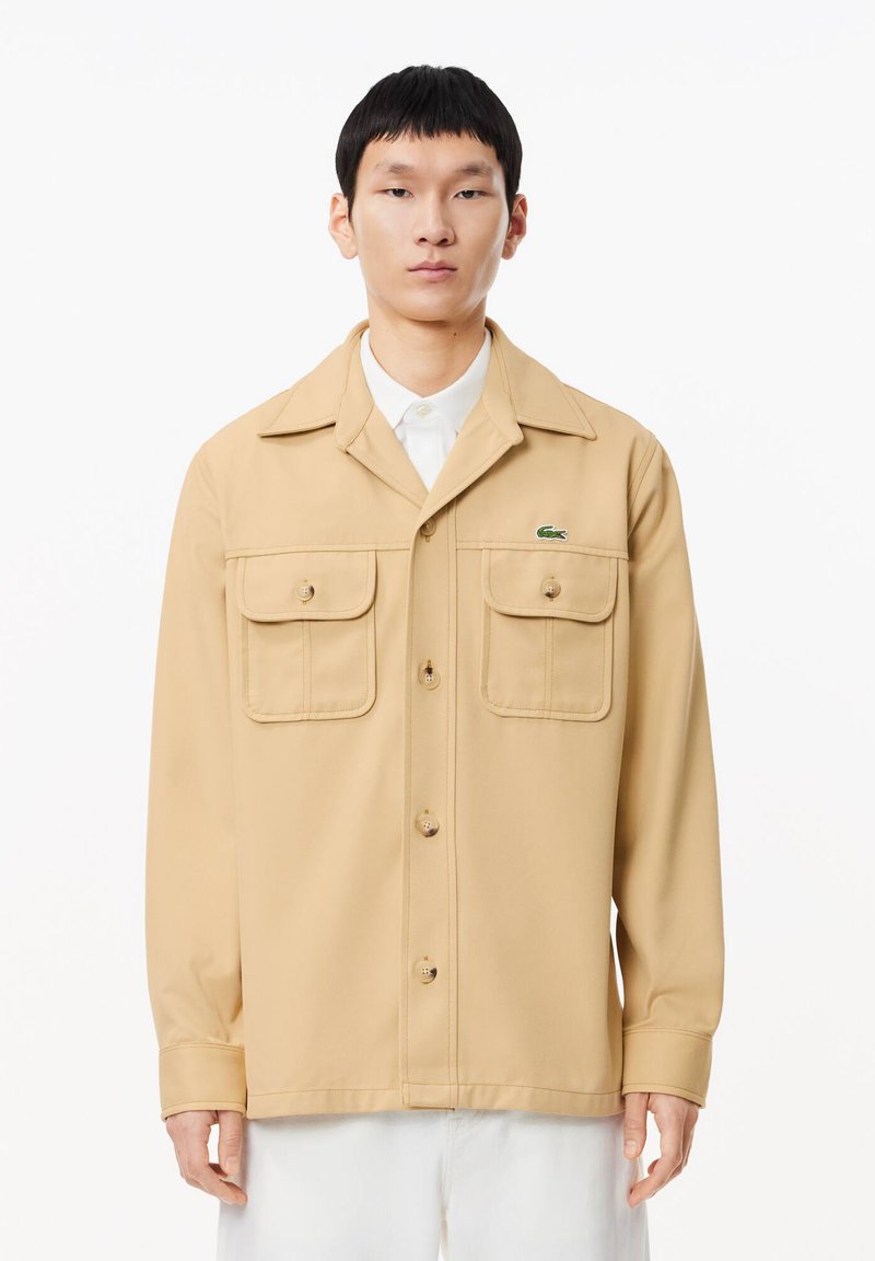 Beige Hemdjacke aus glattem Stoff, mit zwei Brusttaschen, Knopfverschluss und einem kleinen grünen Logo auf der linken Seite.