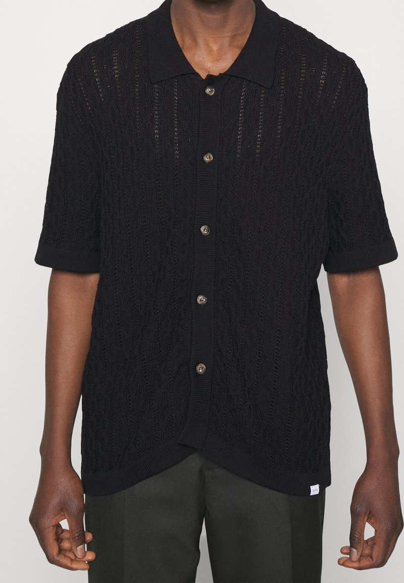 Homme portant une chemise en maille à manches courtes noire avec col et motif en câble texturé, associée à un pantalon foncé.