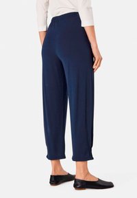 Marineblaue hochgeschnittene Hosen aus weichem Stoff, mit lockerem Sitz und gesäumten Bündchen. Getragen mit schwarzen flachen Schuhen, von hinten gezeigt.