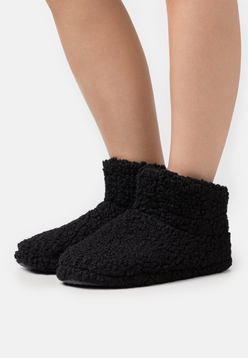 Chaussons d'intérieur en polaire texturée noire portés aux pieds, couvrant les chevilles, avec un design doux, confortable et à haut col sur fond blanc.